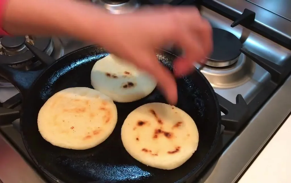 Three arepas browning on a cast-iron skillet over a gas flame, with golden spots forming on the crust