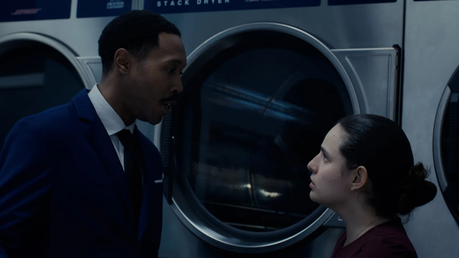 A scene from the 2026 short film In Sanctuary: a man in a suit and a young woman face each other in front of industrial dryers