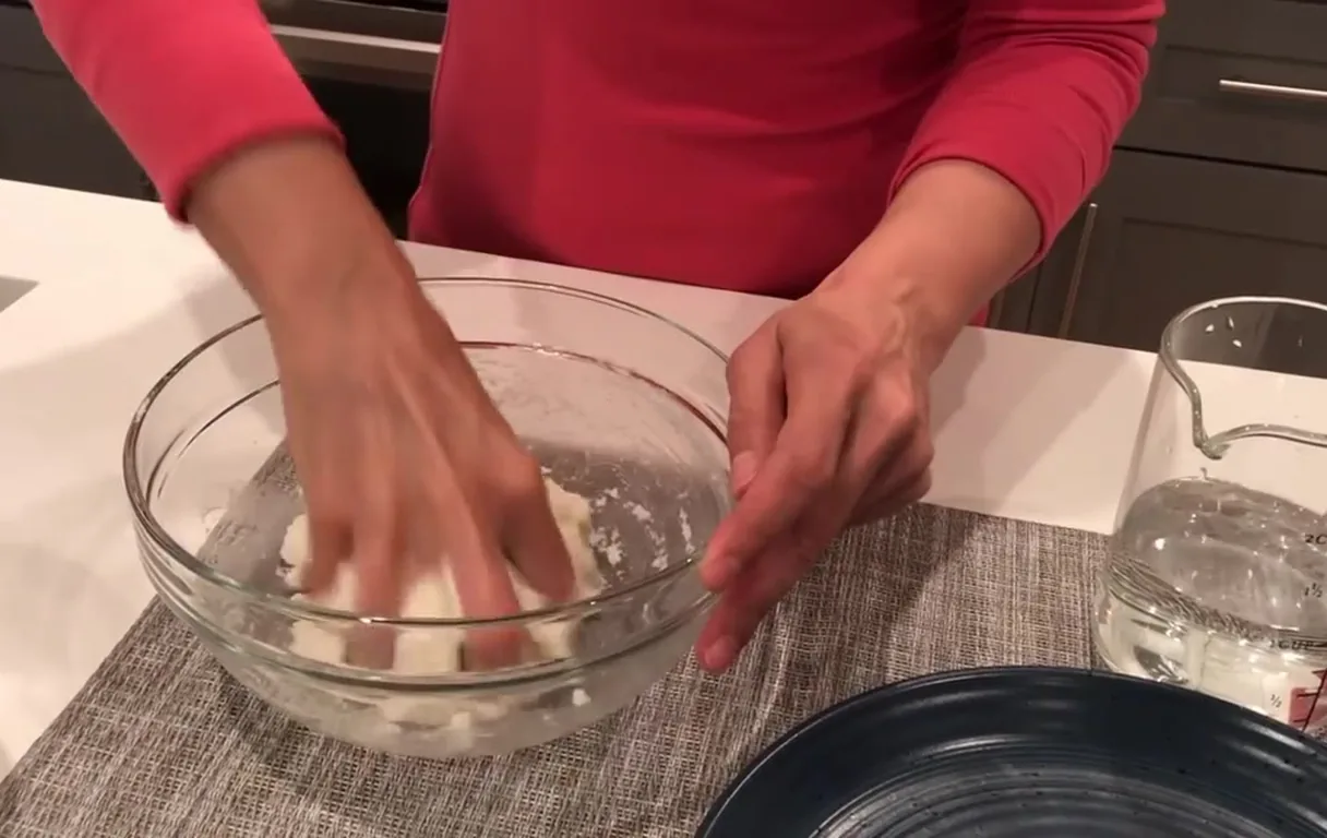 Hands kneading white arepa dough in a glass bowl next to a measuring cup of water